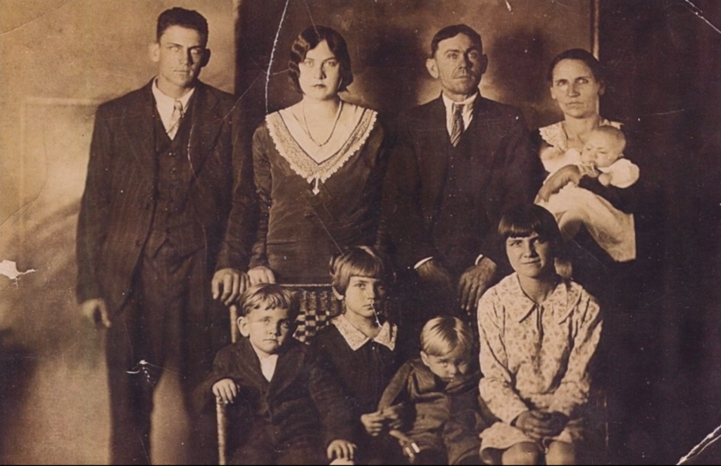 Un portrait de famille vintage aux tons sépia, parfait pour une galerie Elementor, montre trois hommes adultes, deux femmes (l'une tenant un bébé) et quatre enfants vêtus de vêtements du début du XXe siècle, posés à l'intérieur et regardant l'appareil photo.