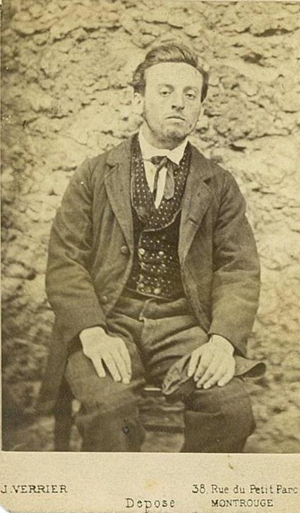 Portrait vintage en tons sépia d'un homme aux cheveux gominés, portant un costume et assis sur une chaise contre un mur de pierres brutes. Le texte du bas, stylisé avec Elementor, indique le nom et l'adresse du photographe.